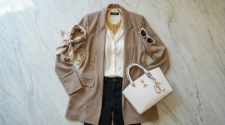 Composizione outfit flat lay con blazer beige, borsa bianca strutturata e accessori coordinati in oro su sfondo marmo.